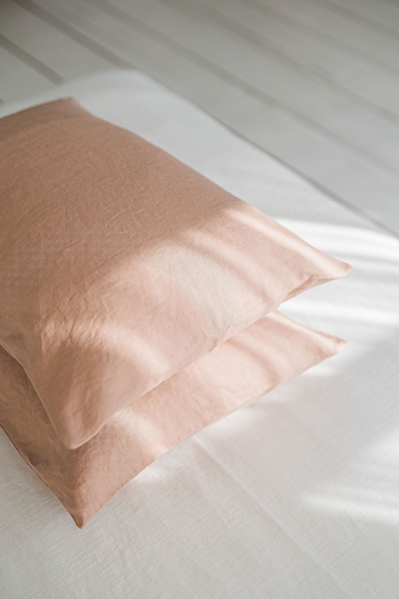 May include: Two peach-colored pillowcases stacked on a white bed sheet. The pillowcases are made of a soft, textured fabric.