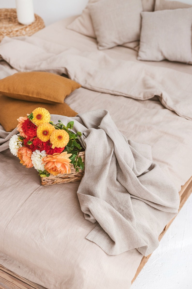 K&ouml;nnte beinhalten: Ein Korb mit orangefarbenen, gelben und roten Blumen steht auf einem Bett mit einer beigefarbenen Bettw&auml;sche aus Leinen und einer Wolldecke.