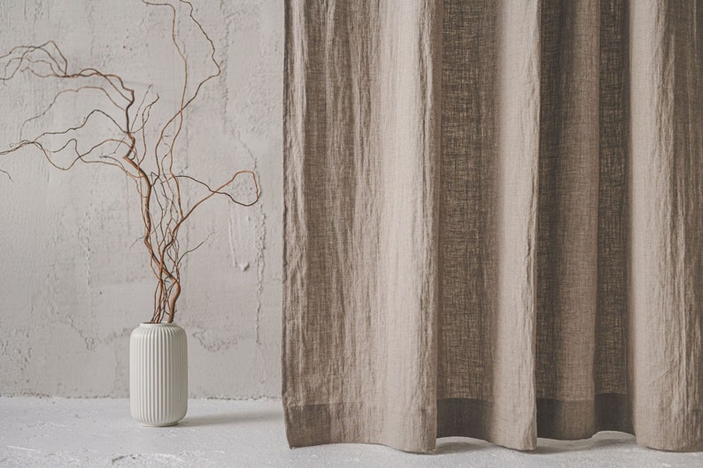 May include: A beige linen curtain with a visible woven texture hangs vertically next to a white cylindrical vase featuring vertical ribbed detailing. Inside the vase, a single bare, twisted brown branch extends upward into the open space.
