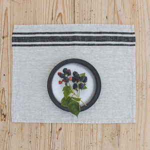 May include: A black ceramic bowl with a sprig of blackberries on a white linen placemat with black stripes. The placemat is on a wooden surface.