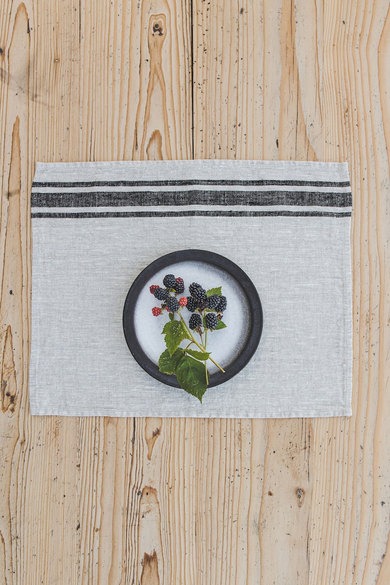 May include: A black ceramic bowl with a sprig of blackberries on a white linen placemat with black stripes. The placemat is on a wooden surface.