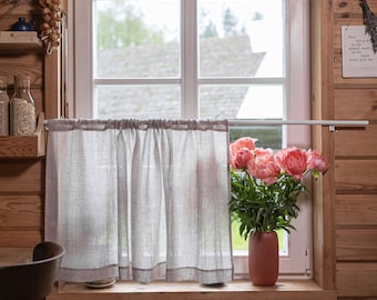 May include: A rustic interior scene featuring a window with a sheer gray curtain and a wooden frame. A vase of pink peonies sits on the windowsill. The room has wooden walls and a hanging bundle of dried herbs.