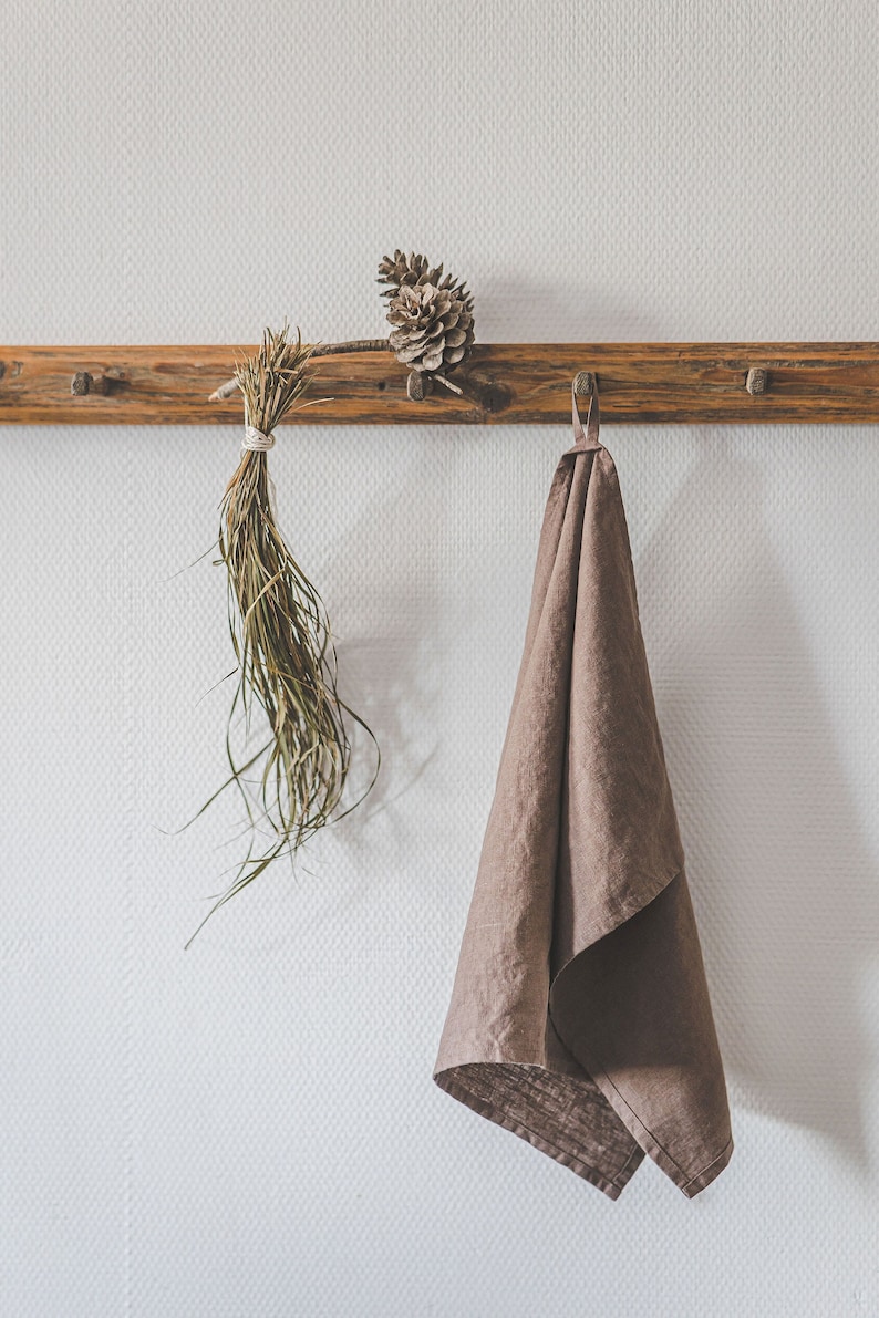 May include: A brown linen kitchen towel hangs from a wooden peg rail. Next to the towel, a bundle of dried green grass and a single pinecone are also suspended from the wooden pegs, creating a display of natural textures and earthy tones.
