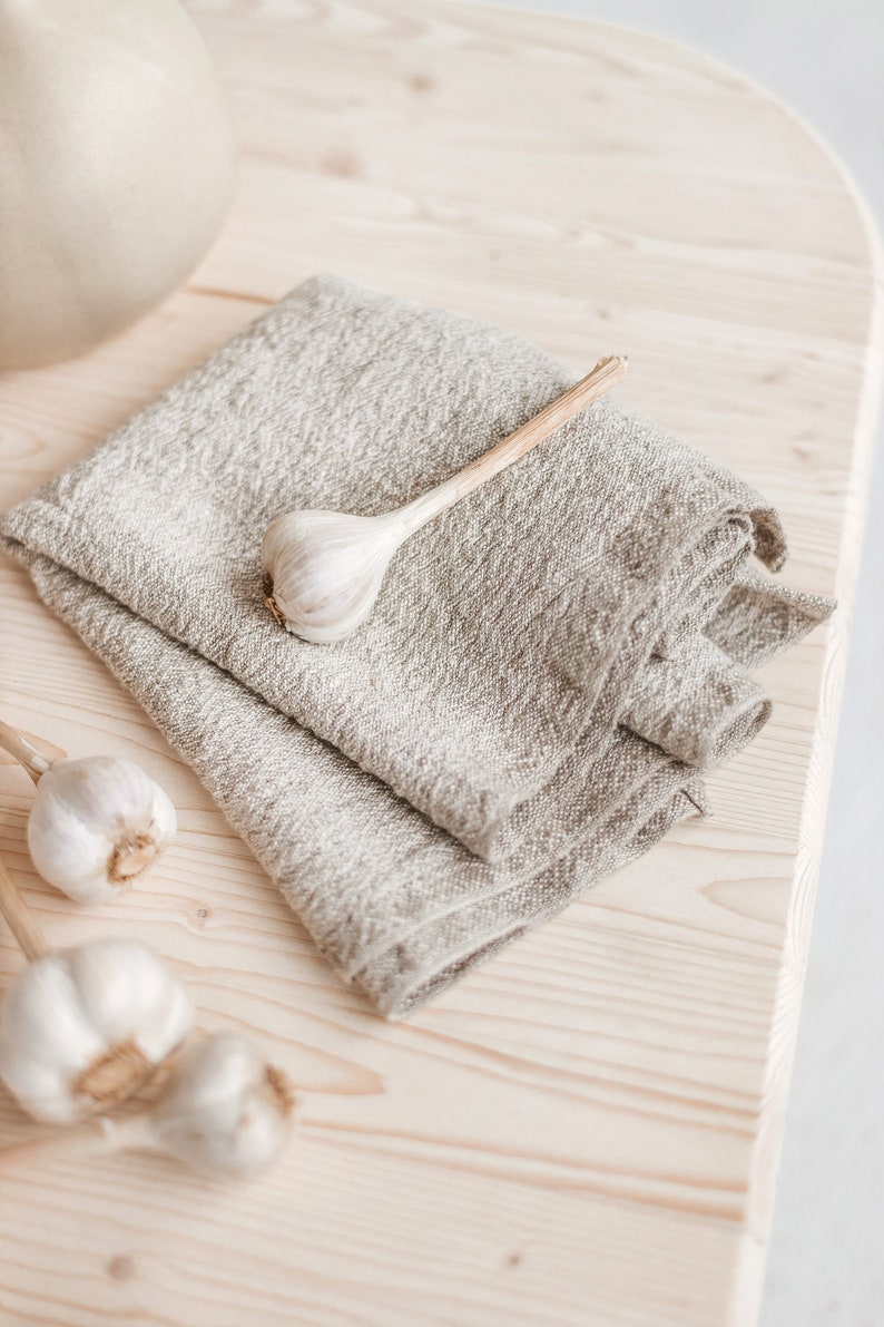 May include: A folded linen kitchen towel on a light wood surface with three whole garlic cloves.