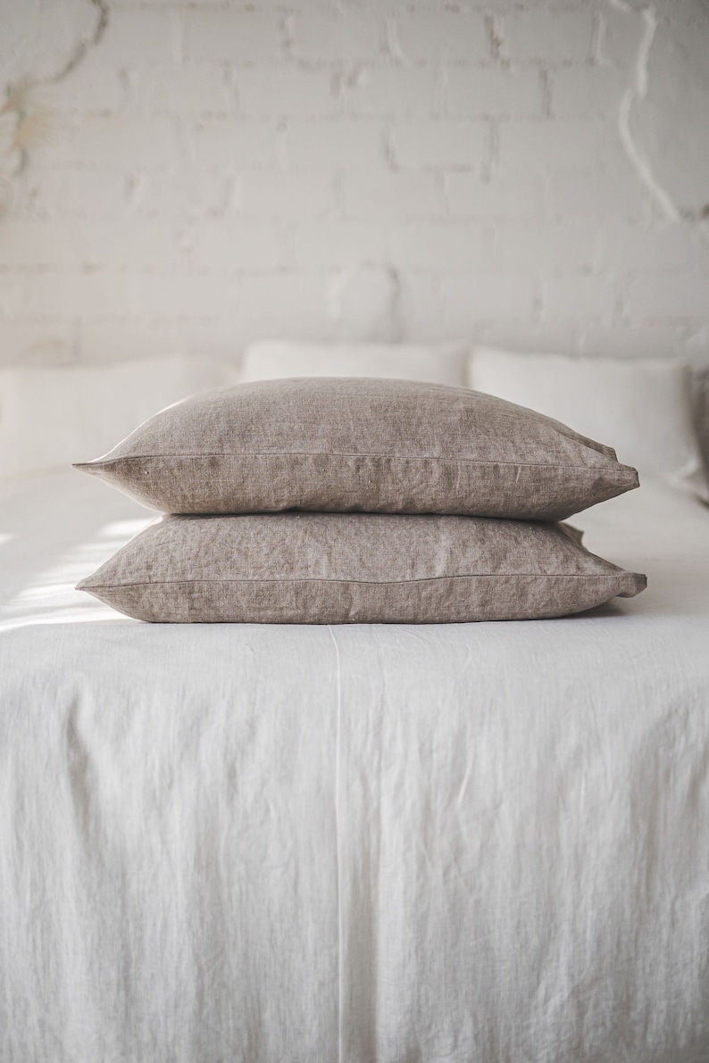 May include: Two brown linen pillowcases stacked on a white bed. The pillowcases are made of a soft, natural fabric and have a subtle texture.
