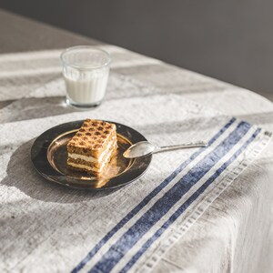 May include: A square piece of layered cake with a light brown crumb topping sits on a silver plate with a silver spoon. The plate is on a linen tablecloth with blue stripes. A glass of milk is in the background.