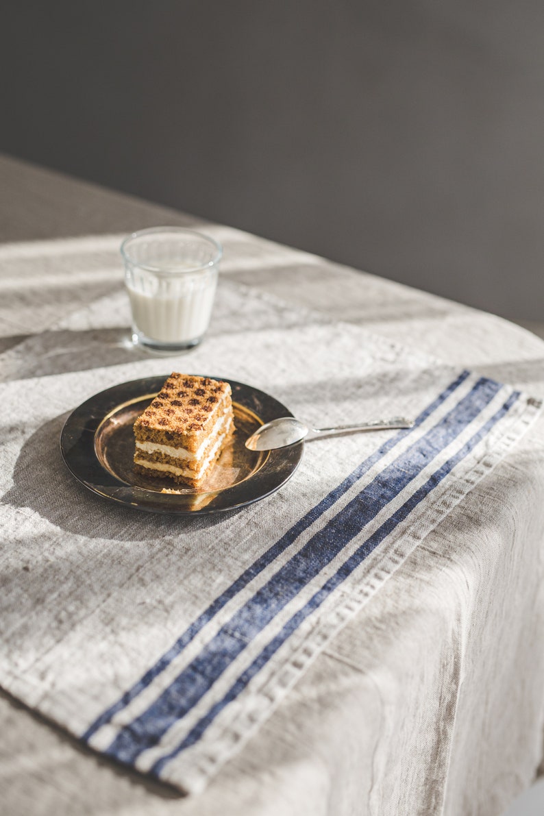May include: A square piece of layered cake with a light brown crumb topping sits on a silver plate with a silver spoon. The plate is on a linen tablecloth with blue stripes. A glass of milk is in the background.
