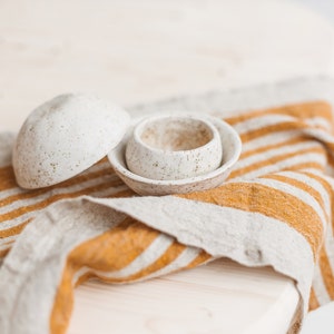 May include: Two white ceramic bowls with a speckled finish, sitting on a striped linen kitchen towel with yellow and white stripes.