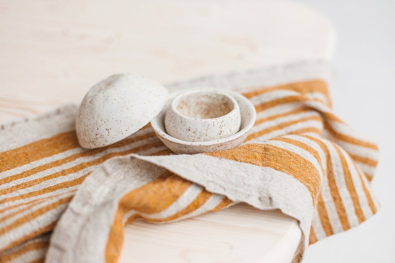 May include: Two white ceramic bowls with a speckled finish, sitting on a striped linen kitchen towel with yellow and white stripes.