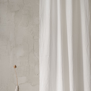 May include: A long, white, textured fabric curtain hangs vertically, featuring subtle folds and a matte finish. Beside the curtain stands a small, irregular, light gray ceramic vase holding a single, dried, brown poppy seed pod on a thin stem.