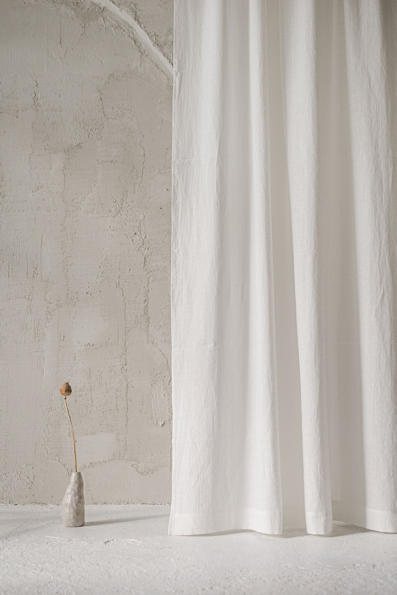 May include: A long, white, textured fabric curtain hangs vertically, featuring subtle folds and a matte finish. Beside the curtain stands a small, irregular, light gray ceramic vase holding a single, dried, brown poppy seed pod on a thin stem.