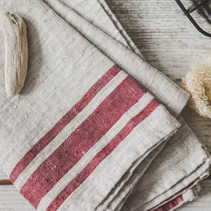 Puede incluir: Una toalla de cocina de lino doblada con dos rayas rojas. La toalla est&aacute; doblada en forma de tri&aacute;ngulo y tiene una est&eacute;tica r&uacute;stica y de granja.