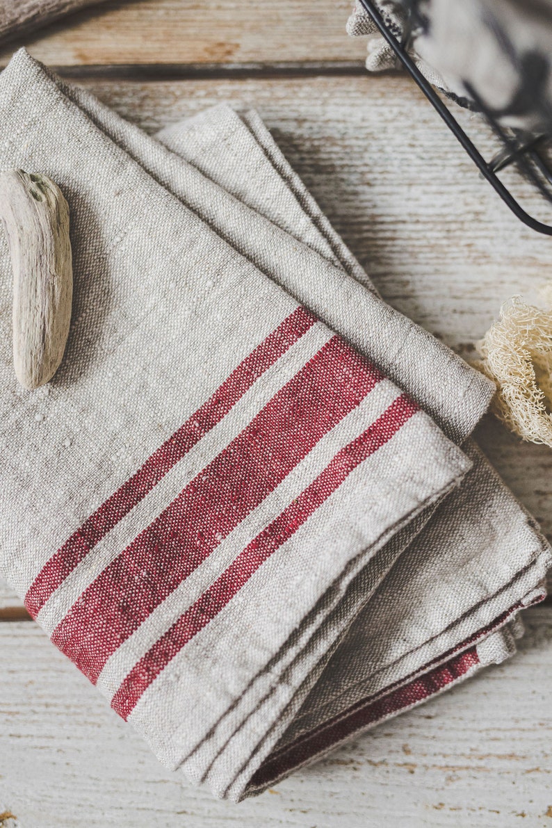 Puede incluir: Una toalla de cocina de lino doblada con dos rayas rojas. La toalla est&aacute; doblada en forma de tri&aacute;ngulo y tiene una est&eacute;tica r&uacute;stica y de granja.