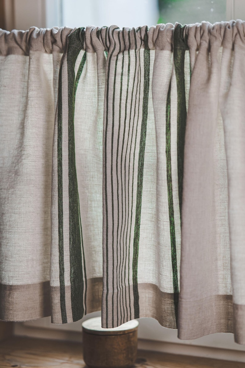 May include: A set of beige and green striped kitchen valances with a gathered top. The valances are made of a linen-like fabric and have a simple, rustic design.