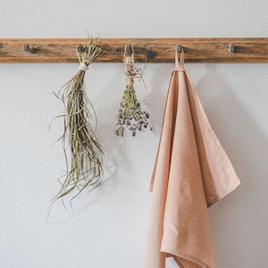 May include: A wooden wall rack with three pegs holds a bundle of long green dried grass, a small bunch of dried purple herbs tied with twine, and a solid light pink kitchen towel hanging from a fabric loop on the rightmost peg.