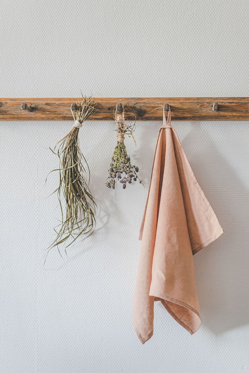 May include: A wooden wall rack with three pegs holds a bundle of long green dried grass, a small bunch of dried purple herbs tied with twine, and a solid light pink kitchen towel hanging from a fabric loop on the rightmost peg.