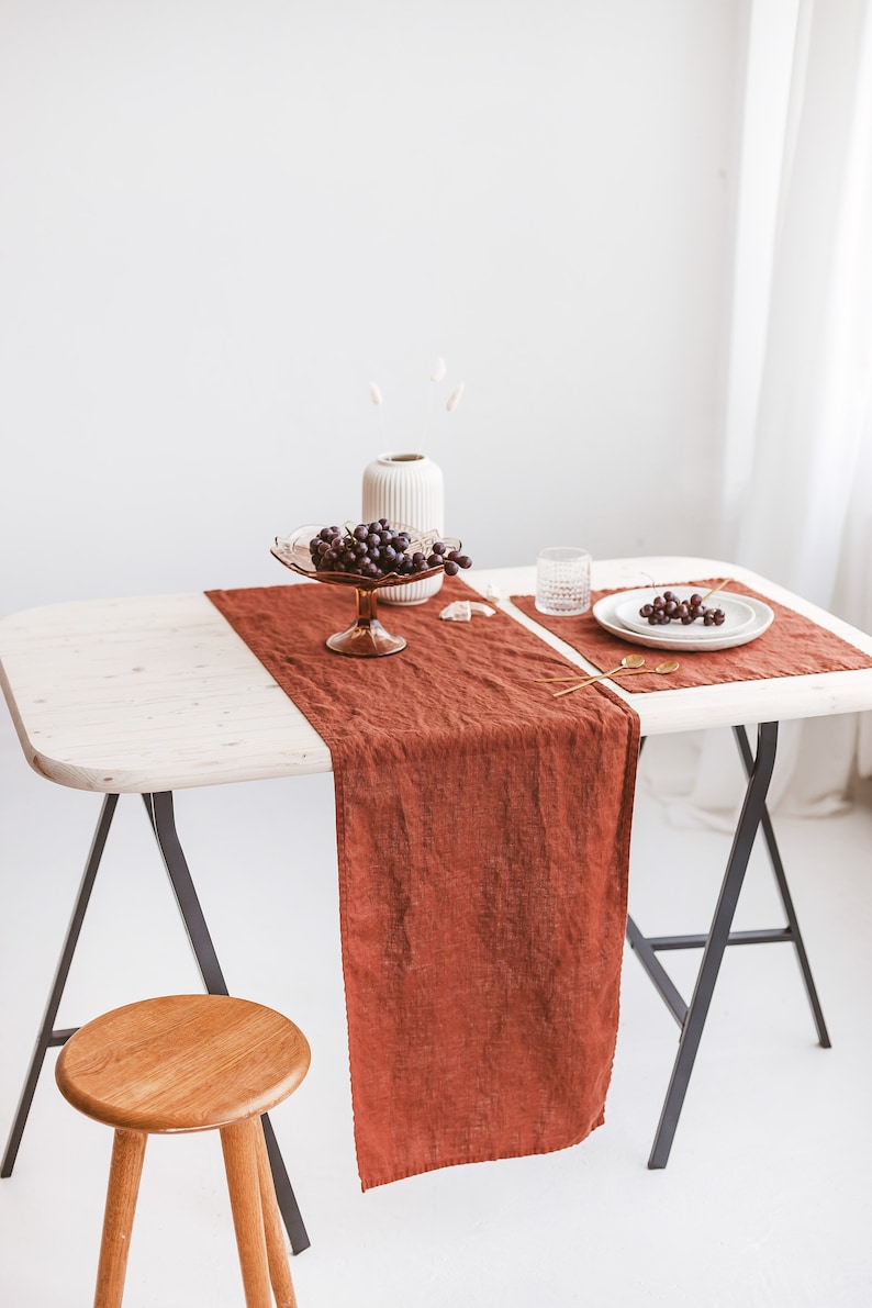 May include: A rustic table setting with a terracotta linen table runner, white plates, and a glass bowl of grapes. The table is set on a pair of black sawhorse legs.