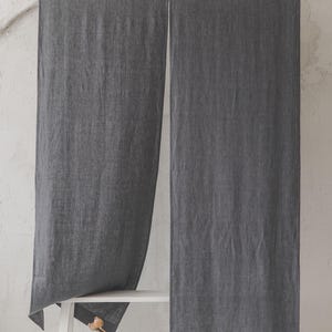 May include: Two charcoal grey linen curtains hanging from a white rod against a white wall. The curtains are long and reach the floor. A white bench is in front of the curtains with a small vase with a dried flower stem.