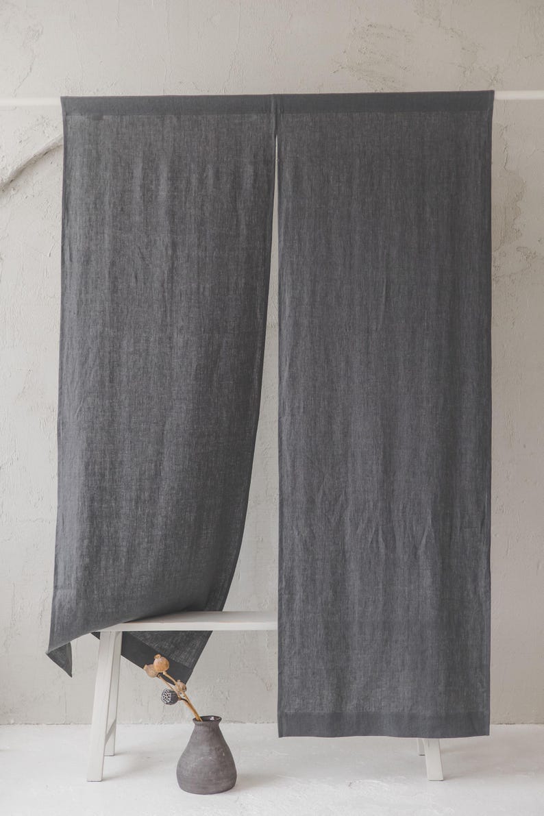 May include: Two charcoal grey linen curtains hanging from a white rod against a white wall. The curtains are long and reach the floor. A white bench is in front of the curtains with a small vase with a dried flower stem.
