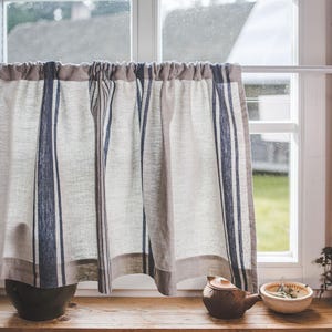 May include: A window with a white frame and a linen curtain with blue stripes. The curtain is pulled to the side, revealing a view of a green lawn. A brown teapot and a white bowl with green leaves are on the windowsill.
