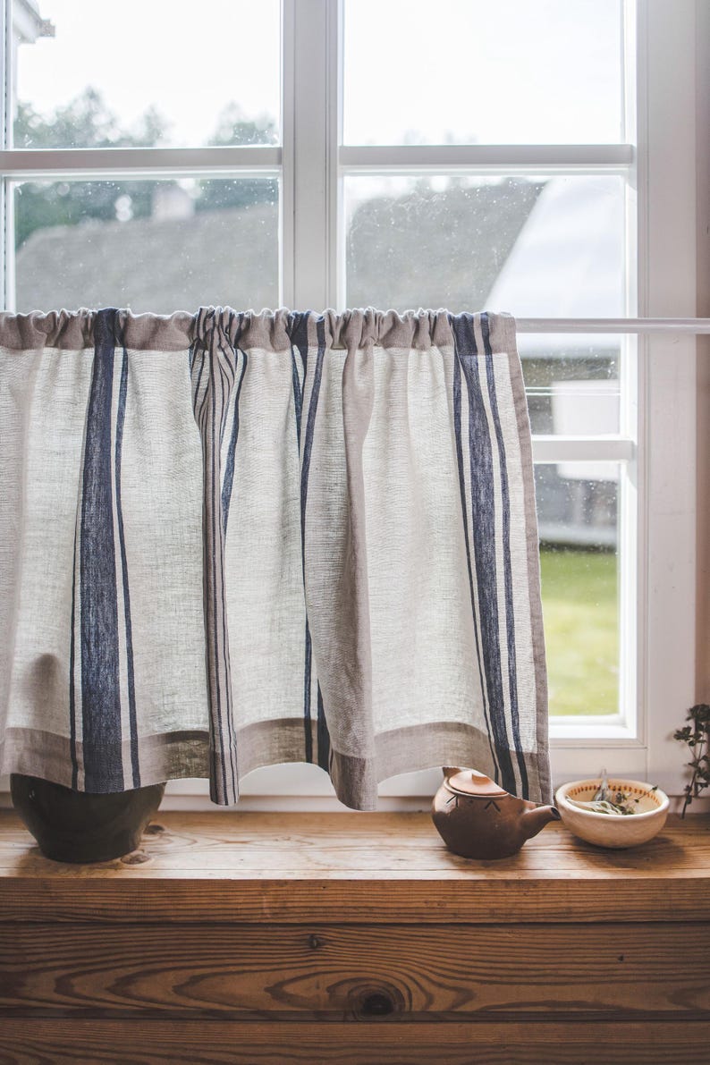 May include: A window with a white frame and a linen curtain with blue stripes. The curtain is pulled to the side, revealing a view of a green lawn. A brown teapot and a white bowl with green leaves are on the windowsill.