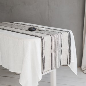 May include: A white linen tablecloth with a black and white striped linen table runner. Two small ceramic bowls are placed on the table runner.