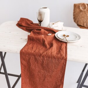 May include: A rust-colored linen table runner on a white wooden table with two gold spoons and a white plate.