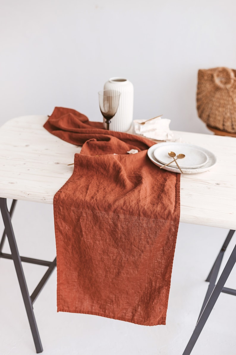 May include: A rust-colored linen table runner on a white wooden table with two gold spoons and a white plate.