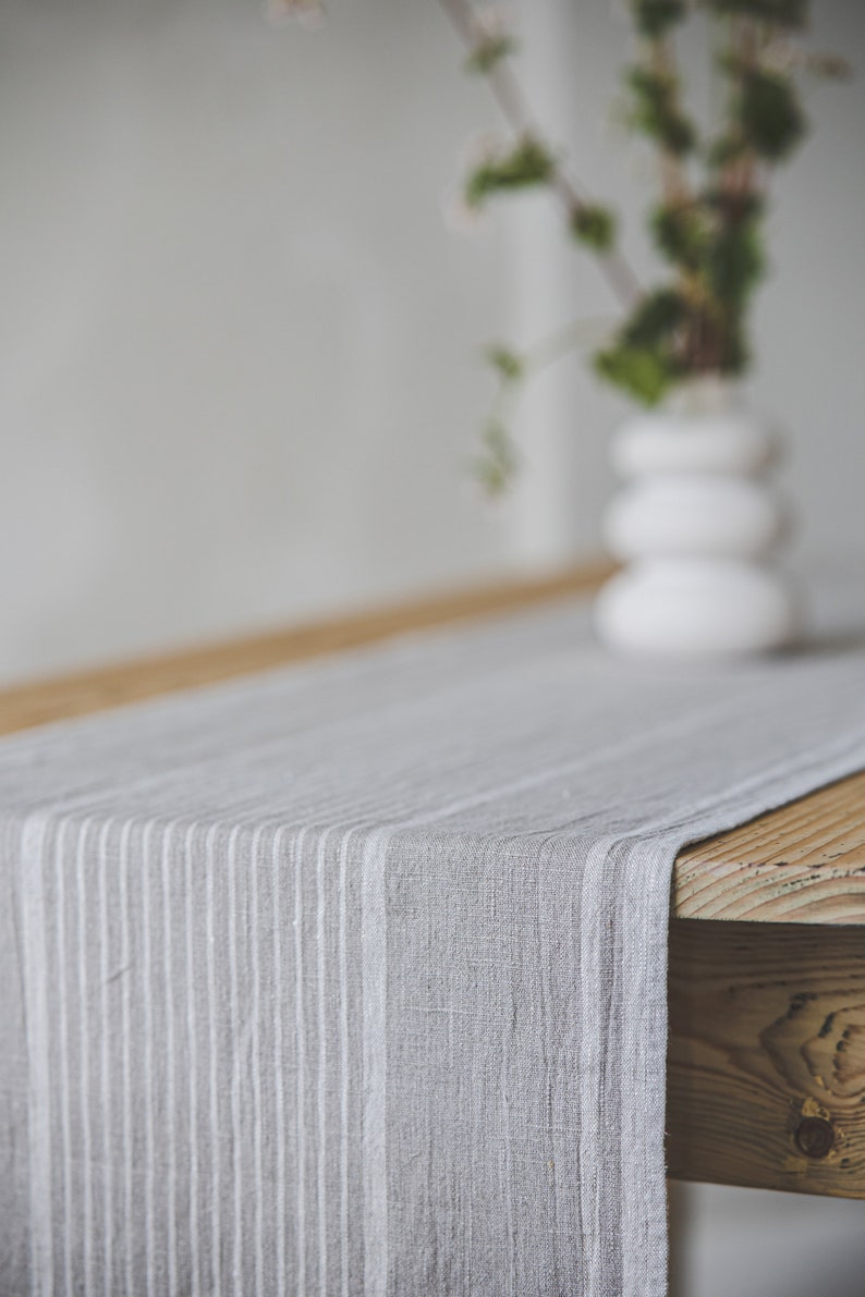 May include: A gray and white striped linen table runner on a wooden table. The runner is made of a soft, natural fabric and has a subtle sheen.
