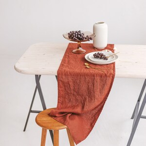 May include: A rustic wooden table with a brown linen table runner, a white vase, a glass bowl with grapes, and two white plates with grapes on a white background.