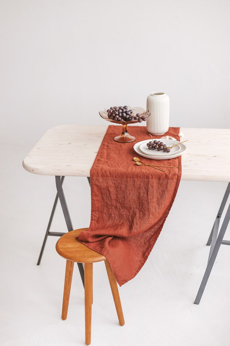 May include: A rustic wooden table with a brown linen table runner, a white vase, a glass bowl with grapes, and two white plates with grapes on a white background.