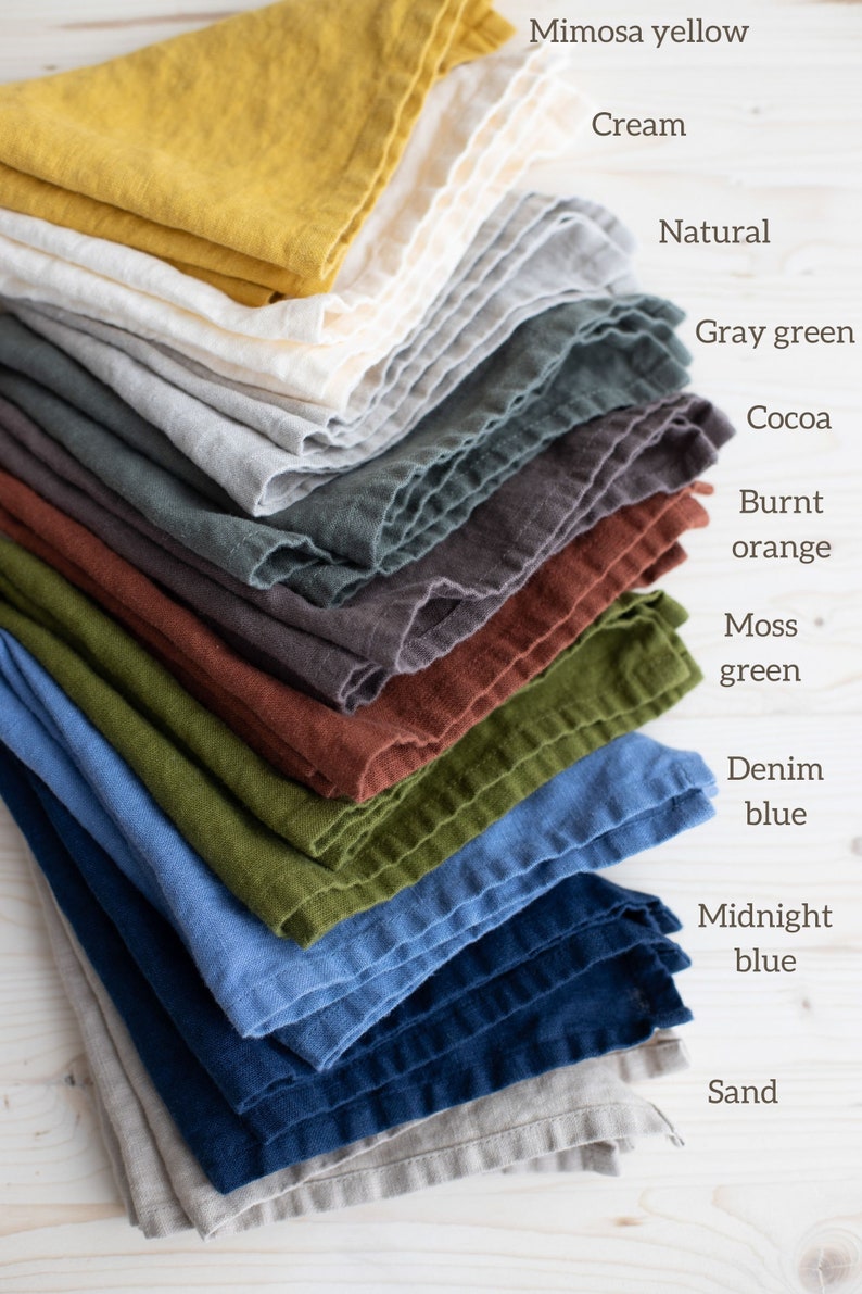 May include: A stack of 12 linen napkins in various colors, including mimosa yellow, cream, natural, gray green, cocoa, burnt orange, moss green, denim blue, midnight blue, and sand.