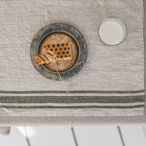 May include: A partially eaten slice of layered cake on a blue and brown ceramic plate with a silver fork. The plate is on a beige linen tablecloth with green stripes. A glass of milk is on the table.