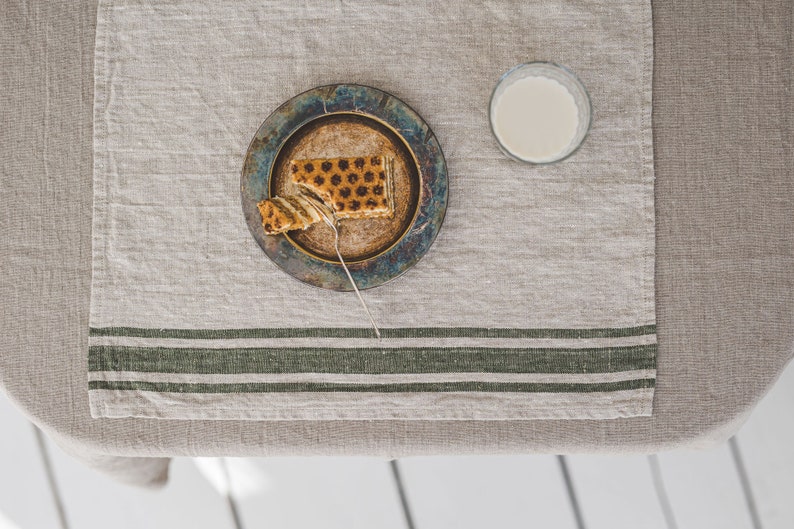 May include: A partially eaten slice of layered cake on a blue and brown ceramic plate with a silver fork. The plate is on a beige linen tablecloth with green stripes. A glass of milk is on the table.