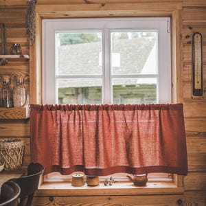 May include: A red linen valance hangs in a window with a wooden frame. The window is in a room with wood paneling on the walls. There are two small ceramic pots on the windowsill.