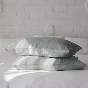 May include: Two sage green linen pillowcases stacked on a white bed. The pillowcases are made of a soft, natural fabric and have a subtle sheen.