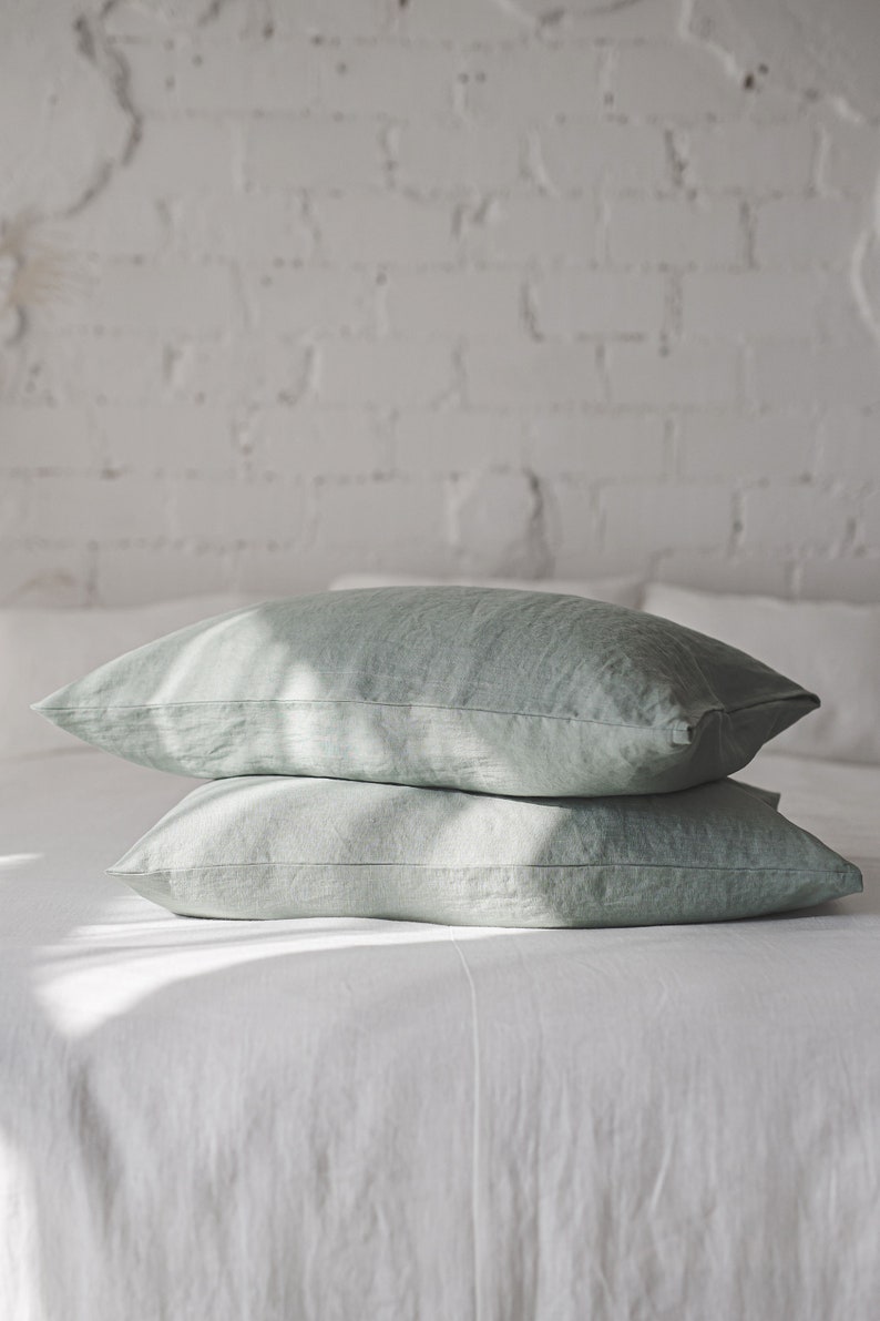 May include: Two sage green linen pillowcases stacked on a white bed. The pillowcases are made of a soft, natural fabric and have a subtle sheen.