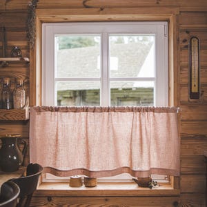 Könnte beinhalten: Ein rosa Leinen-Fensterüberwurf hängt in einem Fenster einer rustikalen Holzhütte. Das Fenster hat einen weißen Rahmen und ist in vier Scheiben unterteilt. Die Wände der Hütte sind aus Holzbrettern.