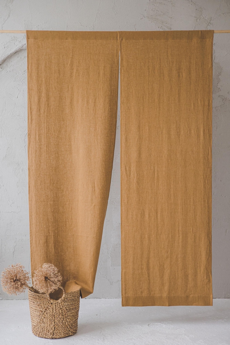 May include: Two panels of light brown linen fabric hanging from a wooden rod. The fabric is textured and has a subtle sheen. The panels are hanging against a white wall. A wicker basket with dried flowers sits in the foreground.