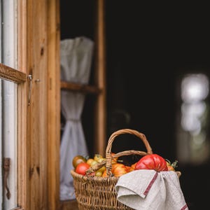 Puede incluir: Una cesta de mimbre llena de tomates rojos y amarillos se encuentra en un alf&eacute;izar de ventana. Un pa&ntilde;o de cocina de lino blanco con rayas rojas cuelga del borde de la cesta.