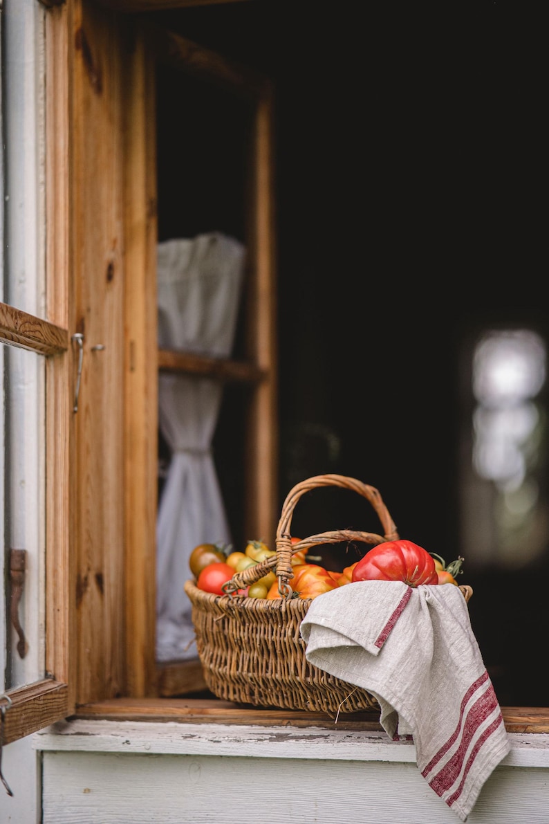 Puede incluir: Una cesta de mimbre llena de tomates rojos y amarillos se encuentra en un alf&eacute;izar de ventana. Un pa&ntilde;o de cocina de lino blanco con rayas rojas cuelga del borde de la cesta.