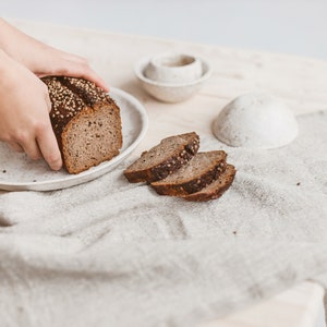 May include: A loaf of dark rye bread with seeds on a white plate, sliced on a white linen tablecloth.