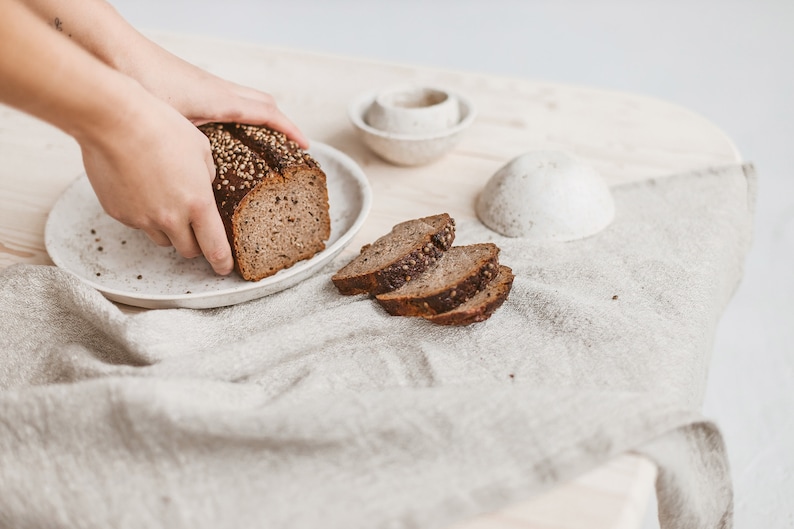 May include: A loaf of dark rye bread with seeds on a white plate, sliced on a white linen tablecloth.