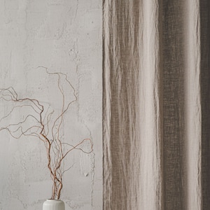 May include: A beige linen curtain hangs vertically next to a white cylindrical vase with vertical ribbing. The vase contains several thin, twisted brown branches that extend upward and outward in a natural, irregular pattern.