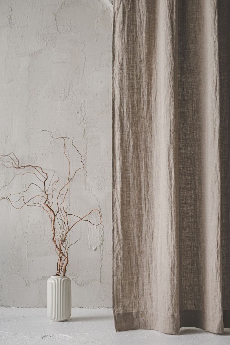May include: A beige linen curtain hangs vertically next to a white cylindrical vase with vertical ribbing. The vase contains several thin, twisted brown branches that extend upward and outward in a natural, irregular pattern.