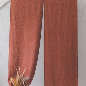 May include: Two rust-coloured linen curtains hanging from a wooden rod against a white wall. A woven basket with dried grasses sits in front of the left curtain.