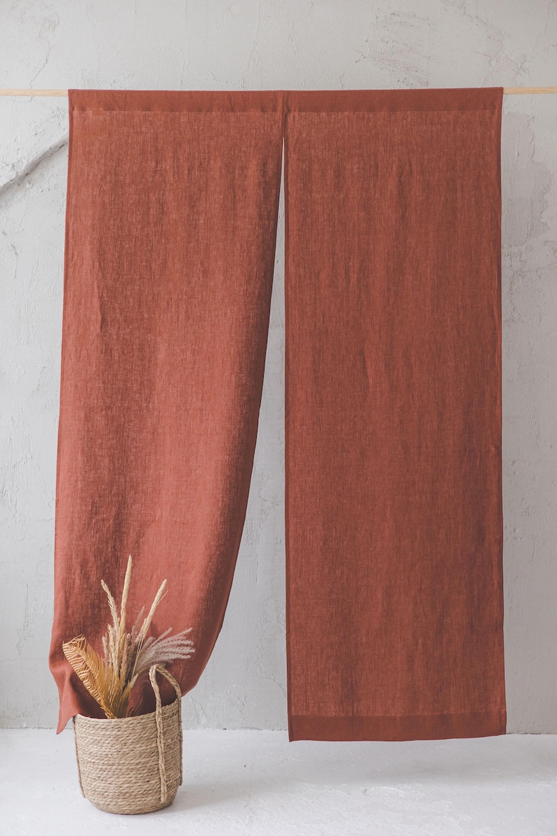 May include: Two rust-coloured linen curtains hanging from a wooden rod against a white wall. A woven basket with dried grasses sits in front of the left curtain.