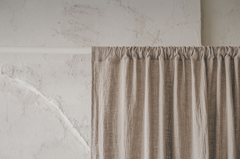 May include: A beige curtain with a visible woven texture hanging from a white rod. The fabric features vertical pleats created by the rod pocket header, resulting in consistent folds that extend downward from the top support structure.