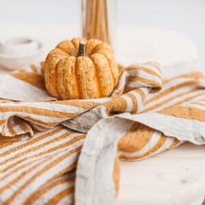 May include: A small, orange pumpkin sits on a striped, beige and brown linen kitchen towel. The towel is folded and draped over a wooden table.
