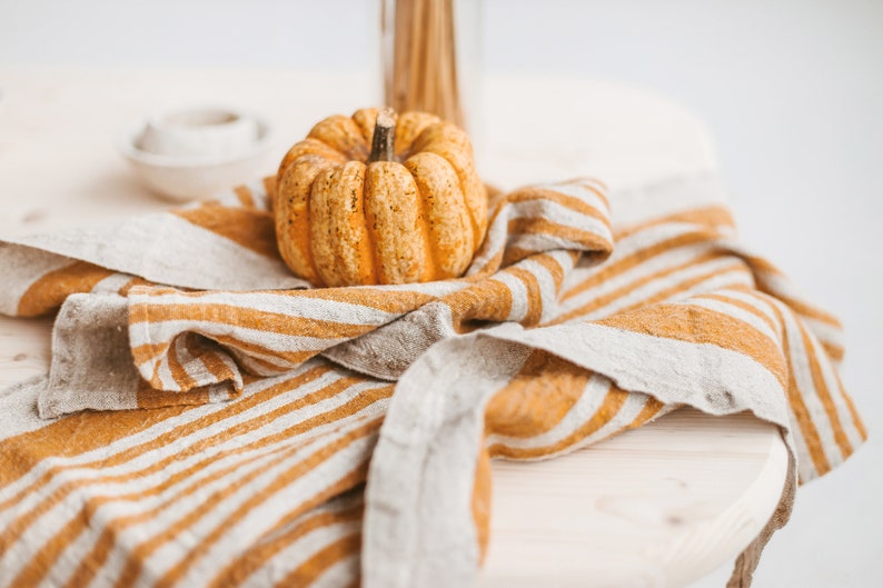 May include: A small, orange pumpkin sits on a striped, beige and brown linen kitchen towel. The towel is folded and draped over a wooden table.